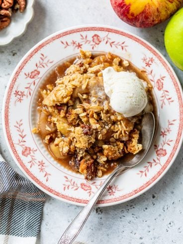 A plate of gluten free apple crisp with a scoop of vanilla ice cream, a spoon, and fresh apples on the side.