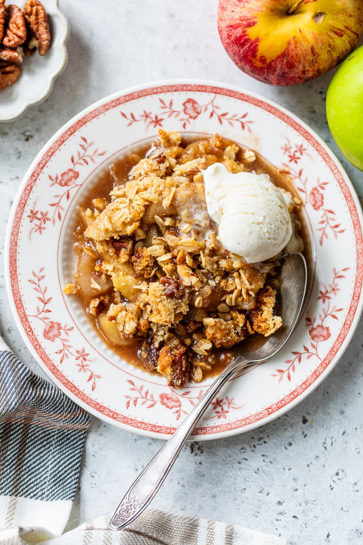 A plate of gluten free apple crisp with a scoop of vanilla ice cream, a spoon, and fresh apples on the side.
