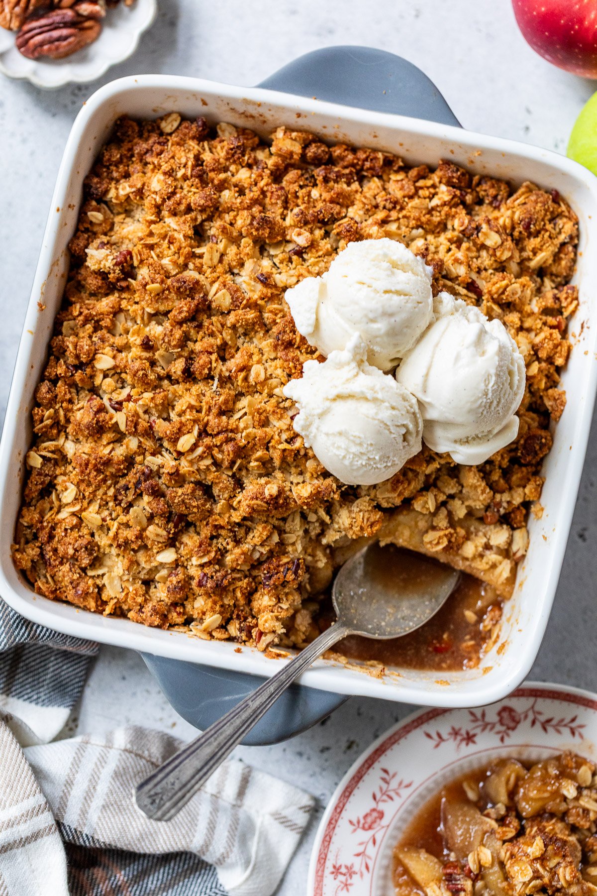 A square dish of gluten free apple crisp topped with three scoops of vanilla ice cream, with a serving spoon and a portion missing.