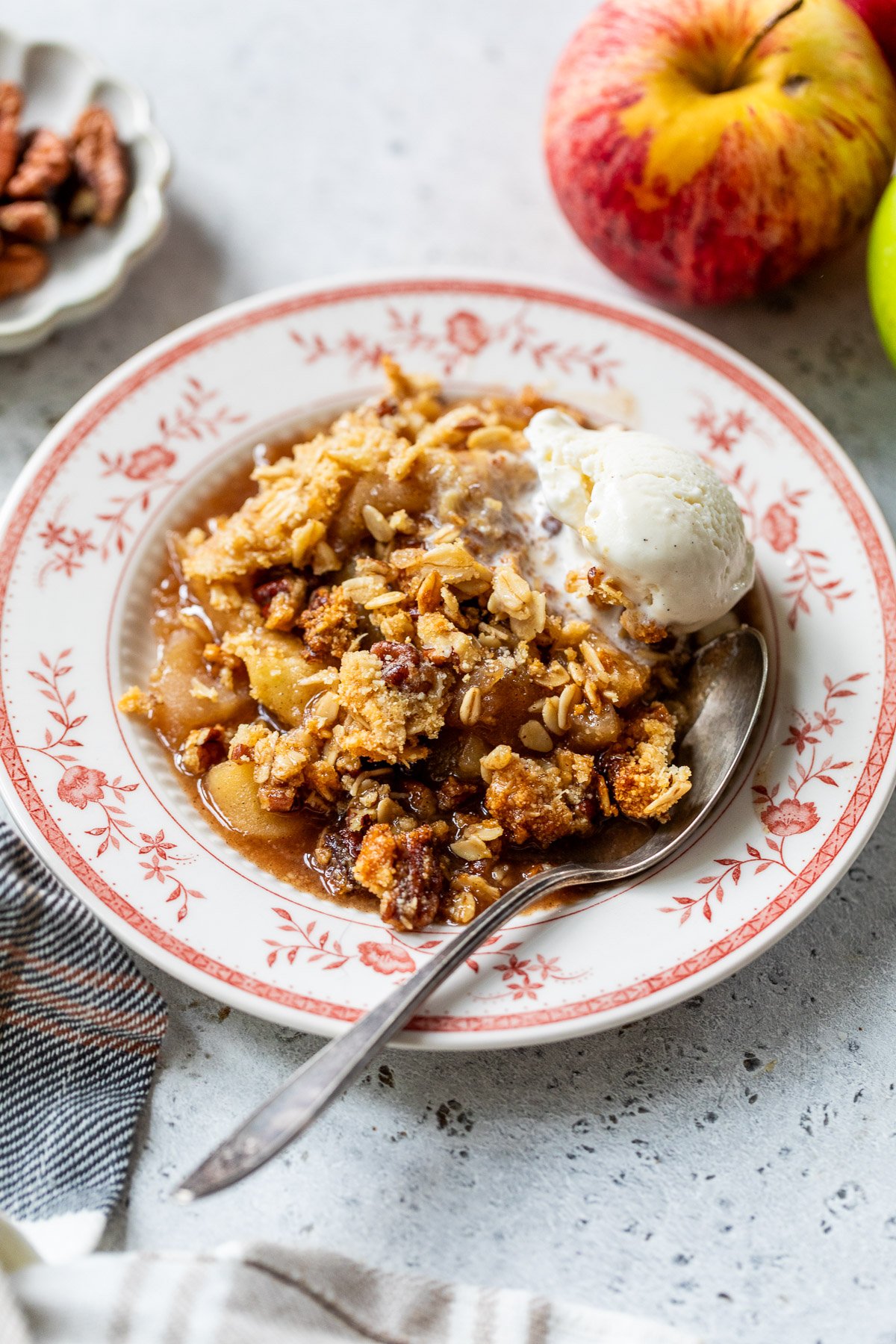 A plate of gluten free apple crisp topped with vanilla ice cream and a spoon, with apples and pecans in the background.