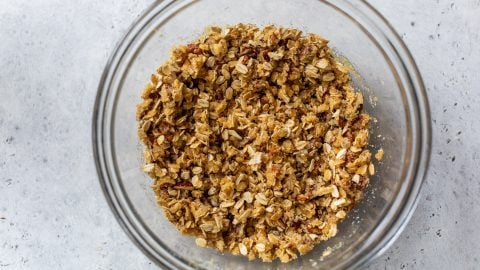 A glass bowl filled with oat and nut crumble topping on a light gray surface.