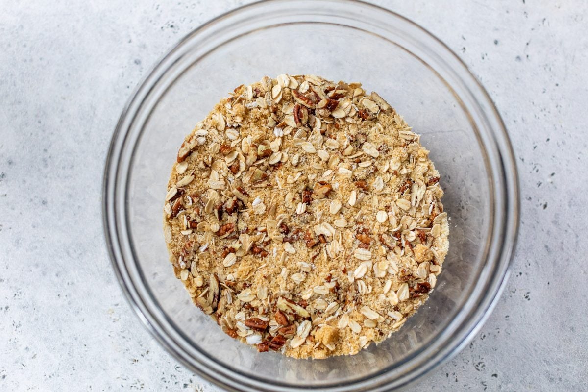 A clear glass bowl filled with oat and nut crumble mixture on a light gray surface.
