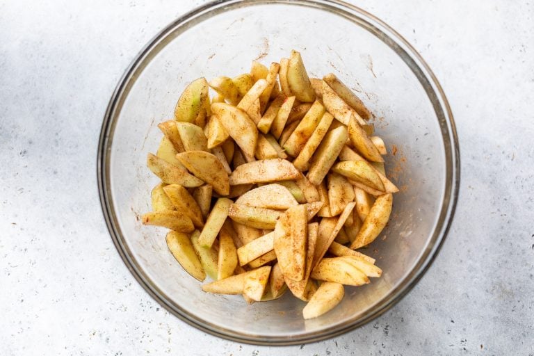 Sliced apples with cinnamon and spices in a clear glass bowl on a light surface.