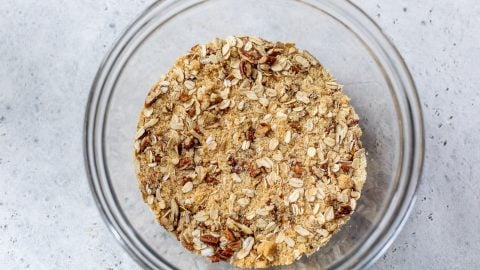A clear glass bowl filled with oat and nut crumble mixture on a light gray surface.