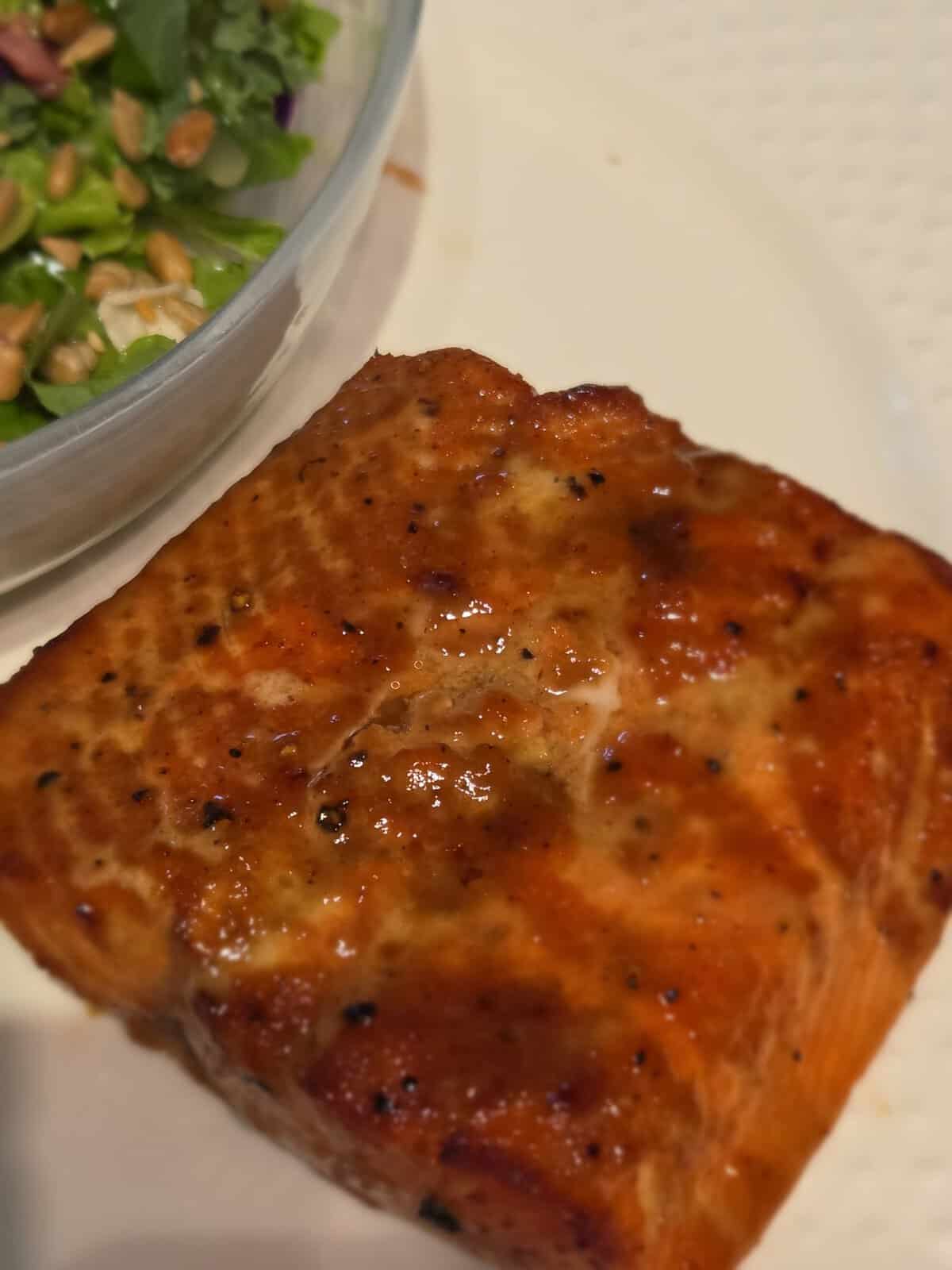 Grilled salmon fillet with a shiny glaze on a white plate next to a bowl of green salad with seeds.