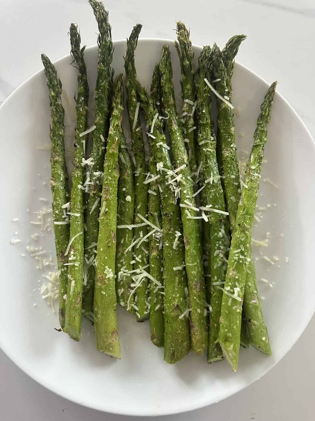 Roasted asparagus spears on a white plate, topped with shredded Parmesan cheese.