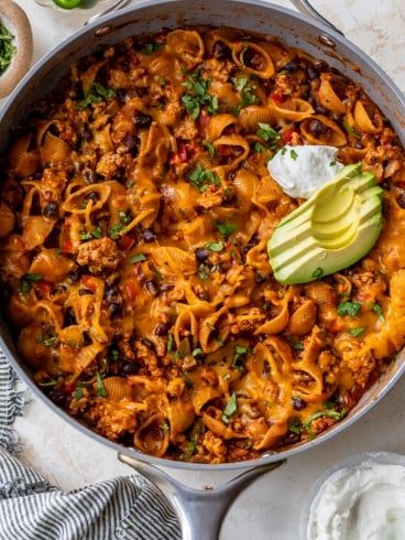 A skillet filled with cheesy taco pasta shells, black beans, and ground meat, topped with sliced avocado, chopped cilantro, and a dollop of sour cream. A striped towel and a bowl of sour cream are nearby.