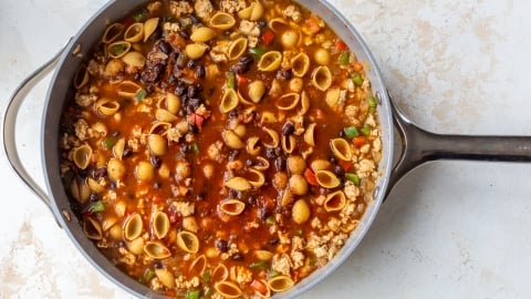 A large pan filled with taco pasta containing small shell pasta, ground meat, black beans, diced tomatoes, and vegetables in a rich, reddish broth sits on a light-colored surface.