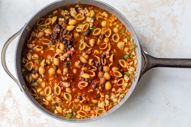A large pan filled with taco pasta containing small shell pasta, ground meat, black beans, diced tomatoes, and vegetables in a rich, reddish broth sits on a light-colored surface.