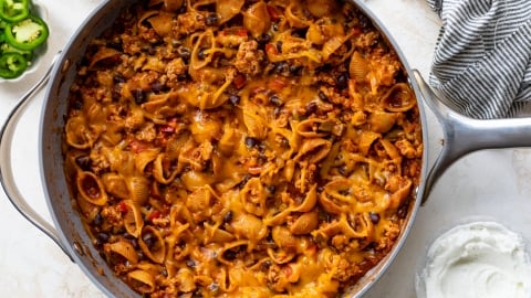 A skillet filled with cheesy taco pasta shells mixed with ground meat, black beans, and melted cheese, next to a bowl of sour cream, sliced jalapeños, and a striped kitchen towel.