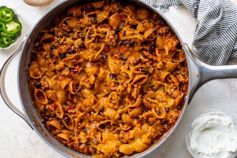 A skillet filled with cheesy taco pasta shells mixed with ground meat, black beans, and melted cheese, next to a bowl of sour cream, sliced jalapeños, and a striped kitchen towel.