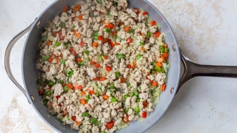 A large skillet filled with cooked ground turkey mixed with diced red and green bell peppers, onions, and carrots on a light, textured surface.