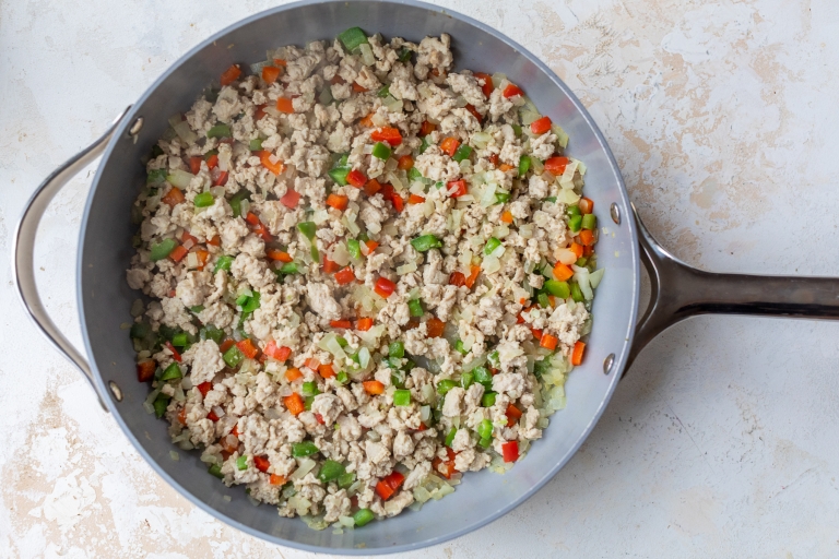 A large skillet filled with cooked ground turkey mixed with diced red and green bell peppers, onions, and carrots on a light, textured surface.