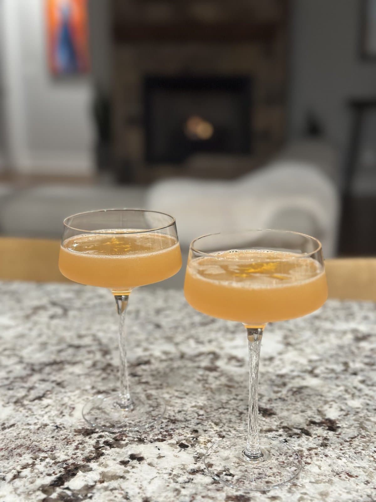 Two coupe glasses filled with a light orange cocktail sit on a speckled countertop, with a cozy living room in the background.