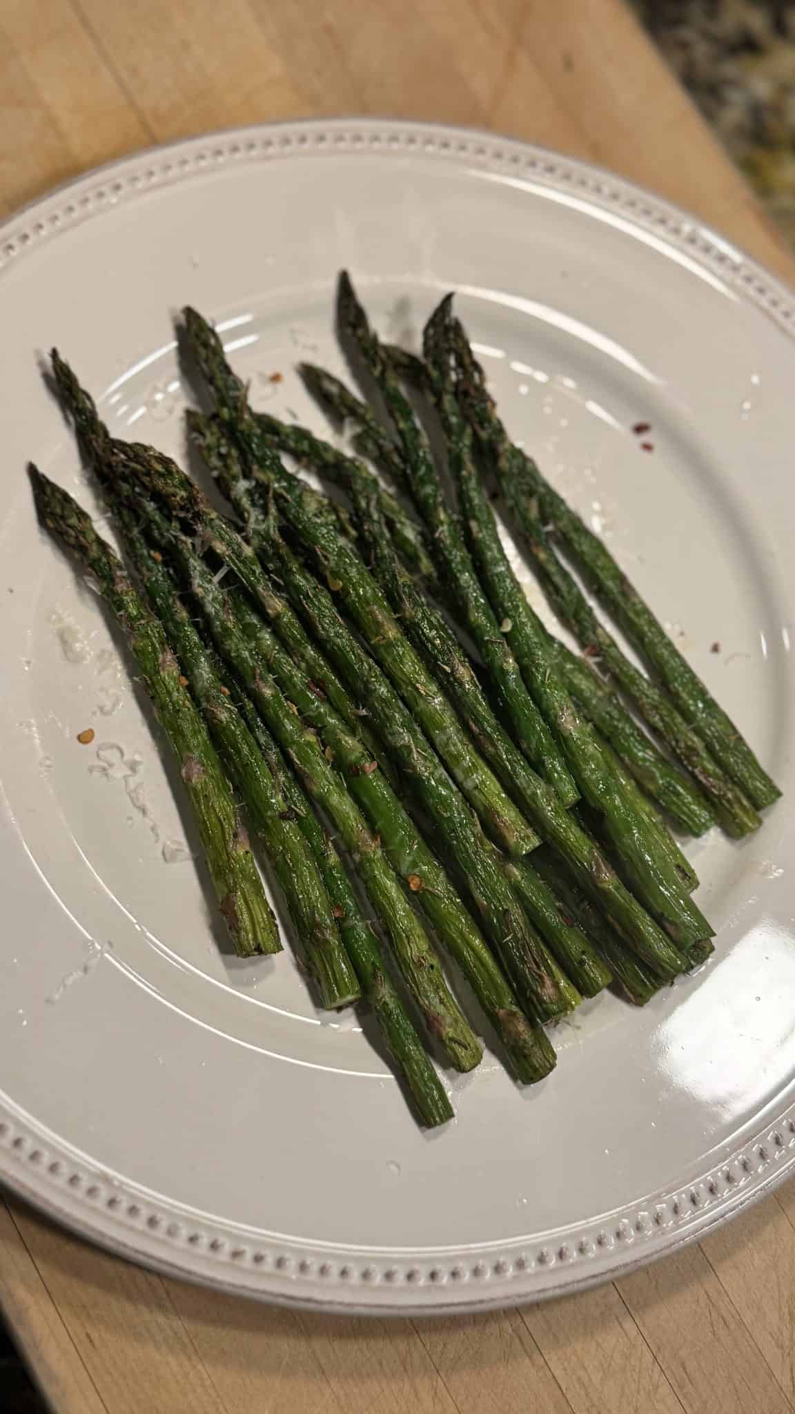 Grilled asparagus spears seasoned and served on a white plate with a beaded edge, placed on a wooden surface.