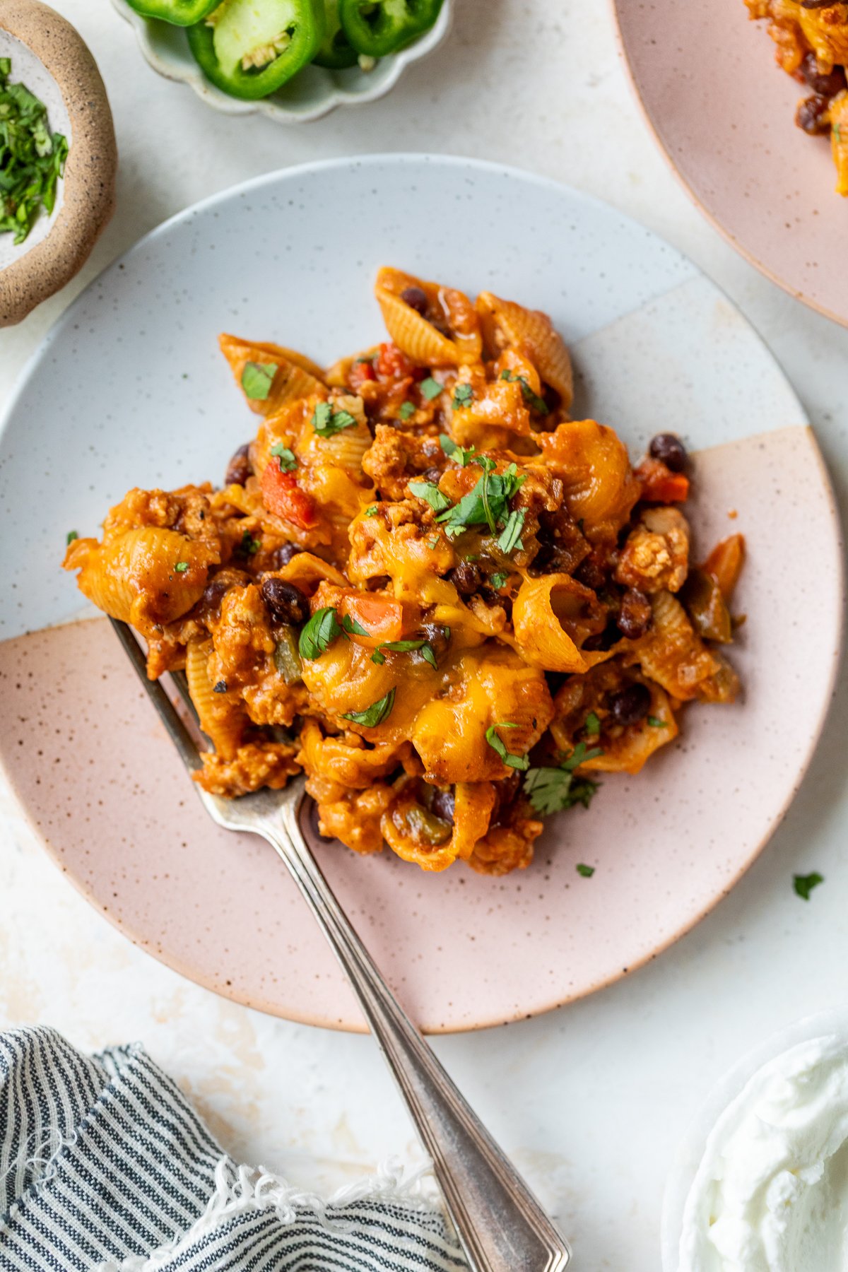 A plate of taco pasta mixed with ground meat, black beans, cheese, and tomato sauce, garnished with chopped herbs. A fork rests on the plate and small bowls with sliced jalapeños and herbs are nearby.
