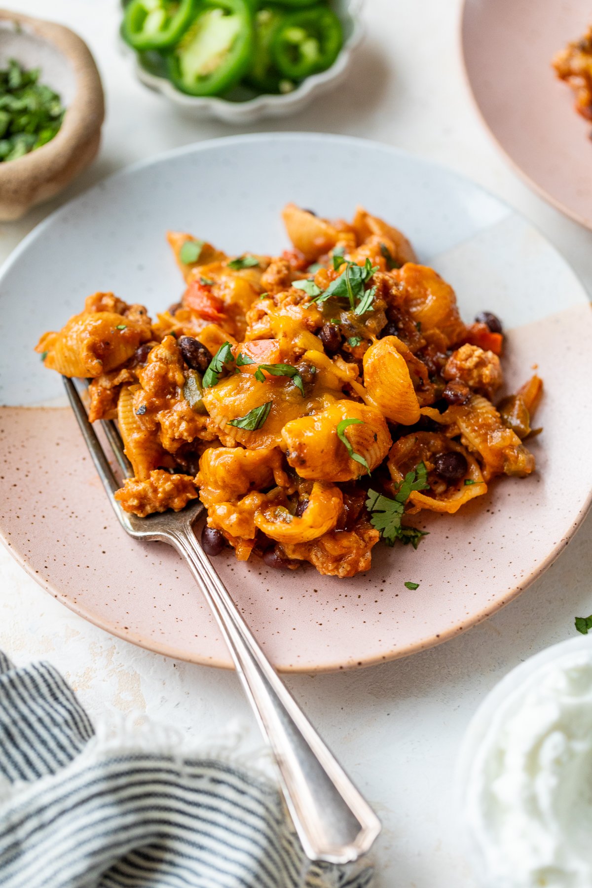 A plate of cheesy taco pasta mixed with ground meat, beans, and garnished with fresh herbs, served with a fork. Sliced jalapeños and other garnishes are visible in the background.