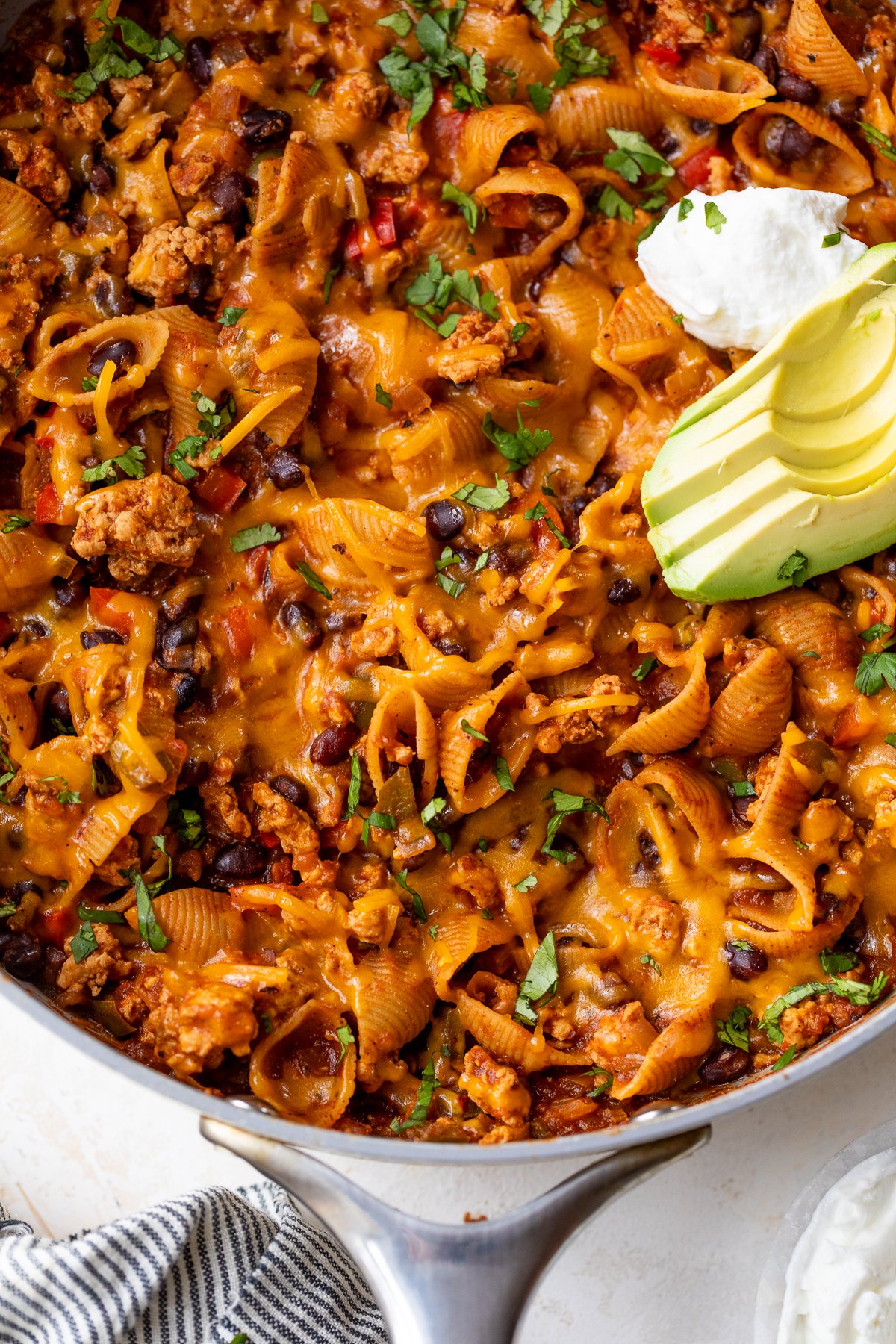 A skillet filled with cheesy taco pasta, black beans, ground meat, and chopped herbs, topped with sliced avocado and a dollop of sour cream.