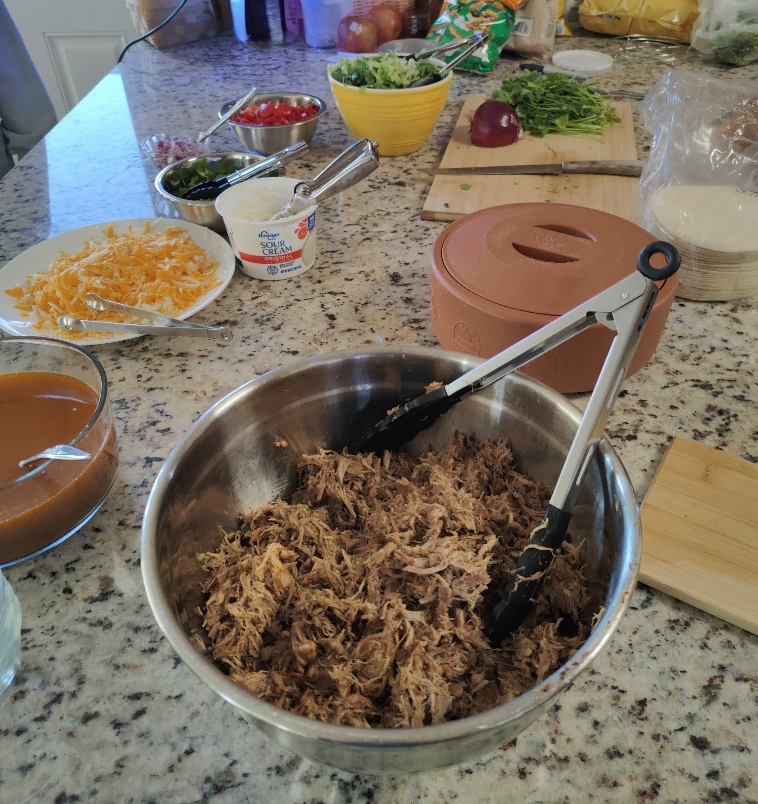 A kitchen counter with shredded meat, cheese, sour cream, salsa, veggies, and toppings for taco or burrito assembly.