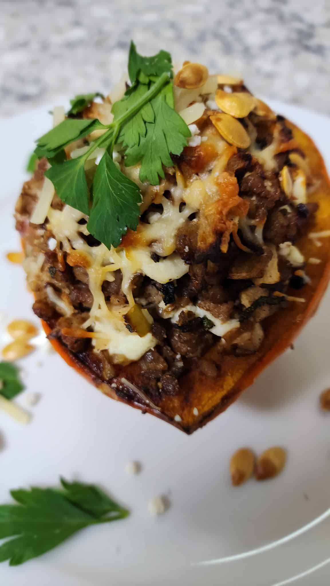 Stuffed butternut squash half topped with melted cheese and parsley on a white plate, garnished with seeds and herbs.