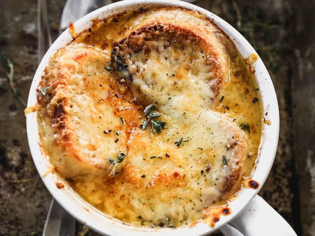 Two white mugs filled with French onion soup, topped with toasted bread and melted cheese, on a rustic tray.