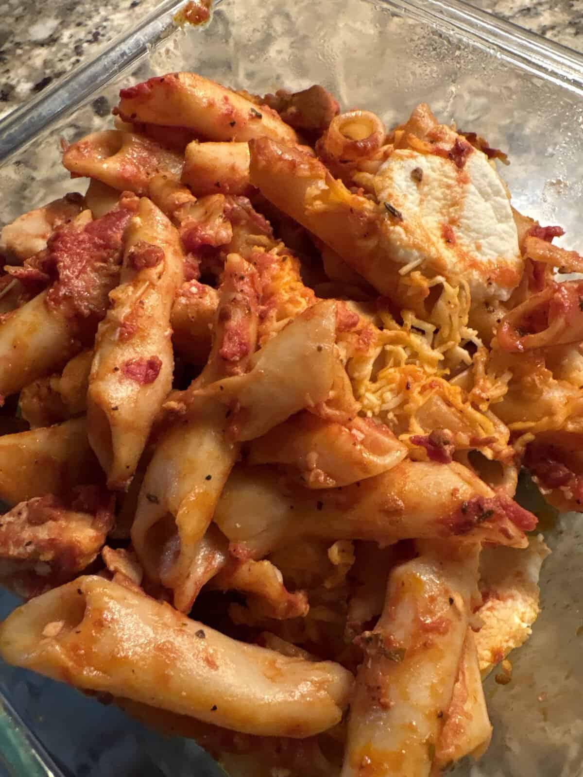 Baked ziti pasta with tomato sauce, melted cheese, and visible herbs in a glass dish on a countertop.