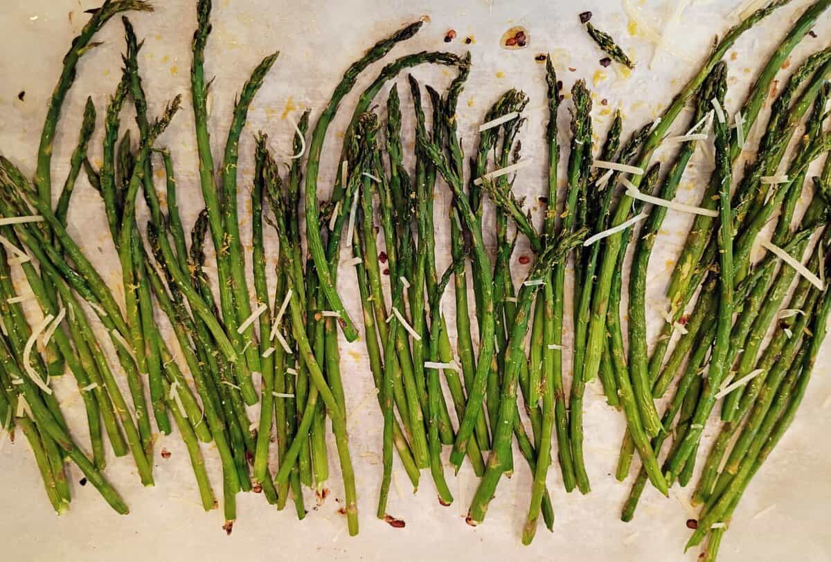 Roasted asparagus spears spread out on a baking sheet, topped with shredded cheese and seasonings.