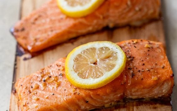 Two cooked salmon fillets on a cedar plank, each topped with a slice of lemon and sprinkled with herbs and pepper.