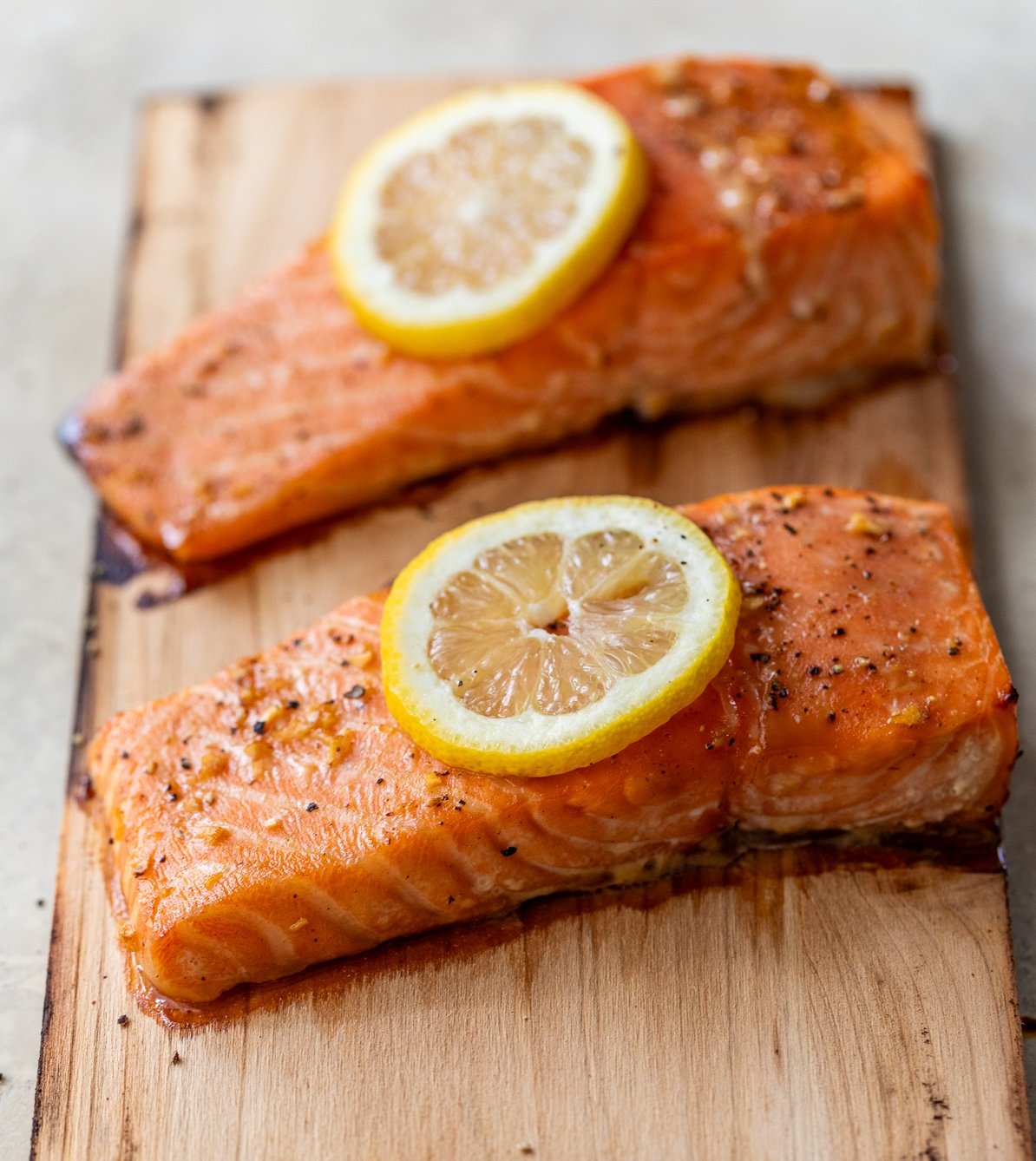Two cooked salmon fillets on a cedar plank, each topped with a slice of lemon and sprinkled with herbs and pepper.