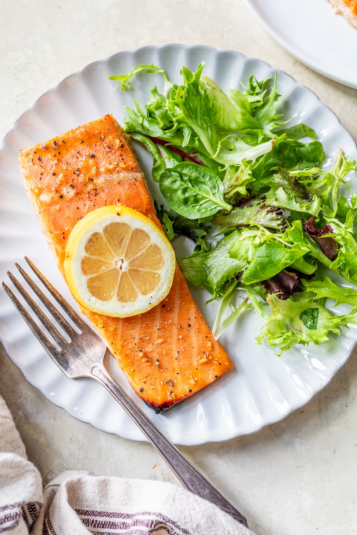 A plate with a piece of cedar plank salmon topped with a lemon slice, accompanied by a fresh green salad. A fork sits beside the food on the white plate.
