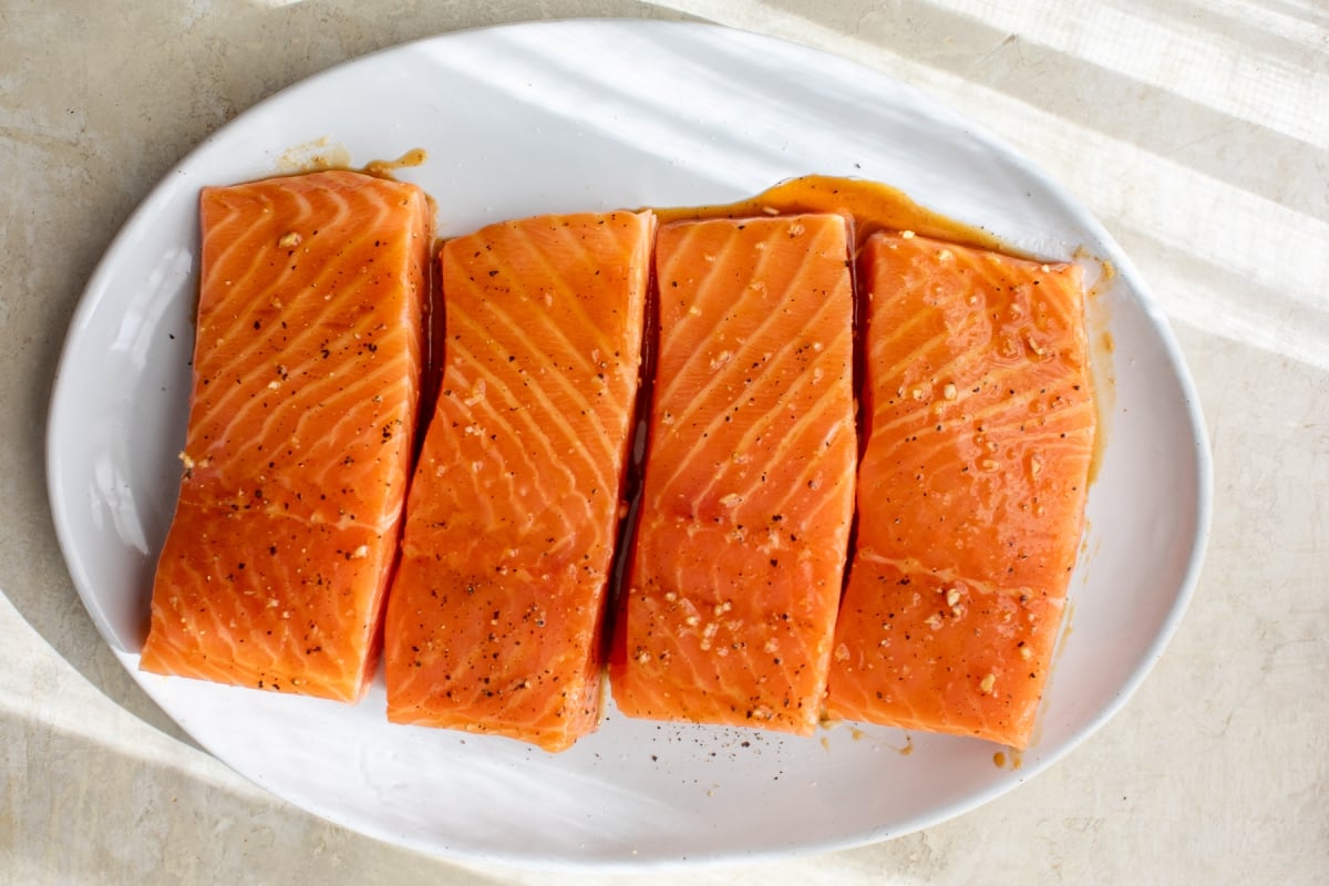 Four raw salmon fillets, seasoned with salt and pepper, are arranged in a row on a white oval plate, with sunlight casting shadows across the surface.