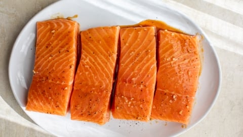 Four raw salmon fillets, seasoned with salt and pepper, are arranged in a row on a white oval plate, with sunlight casting shadows across the surface.