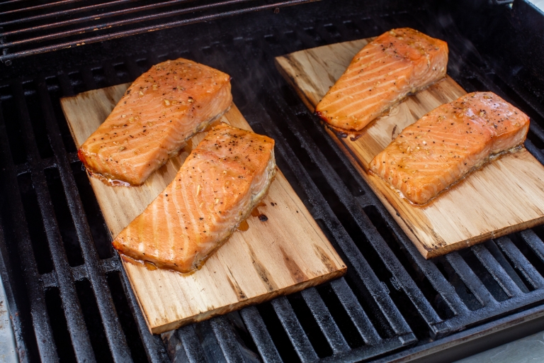 Four seasoned salmon fillets are cooking on cedar planks placed over the grates of a grill. The fish is lightly browned at the edges, and the grill is emitting a bit of steam or smoke.