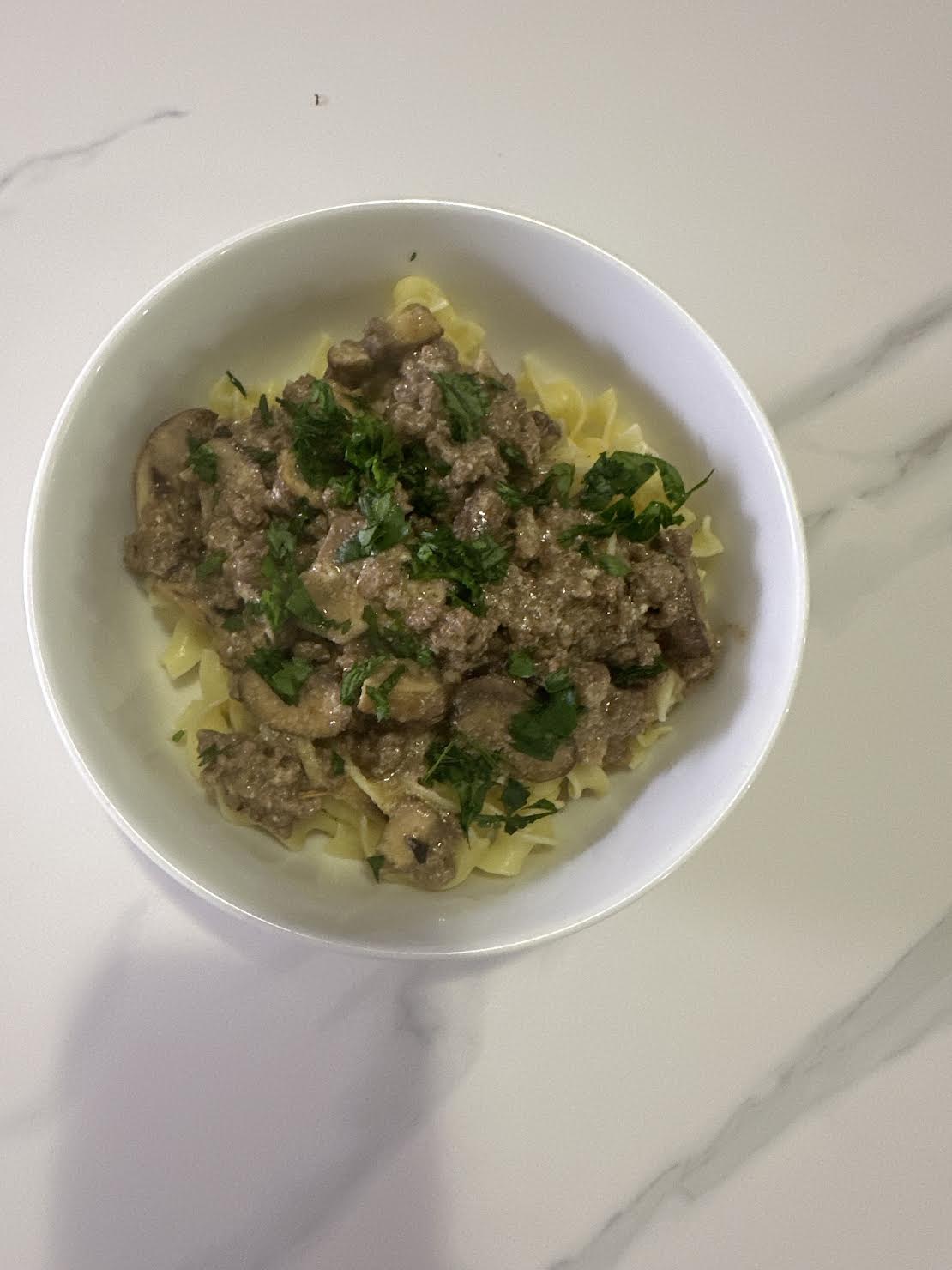 A bowl of pasta topped with creamy beef and mushroom sauce, garnished with chopped parsley on a white marble surface.