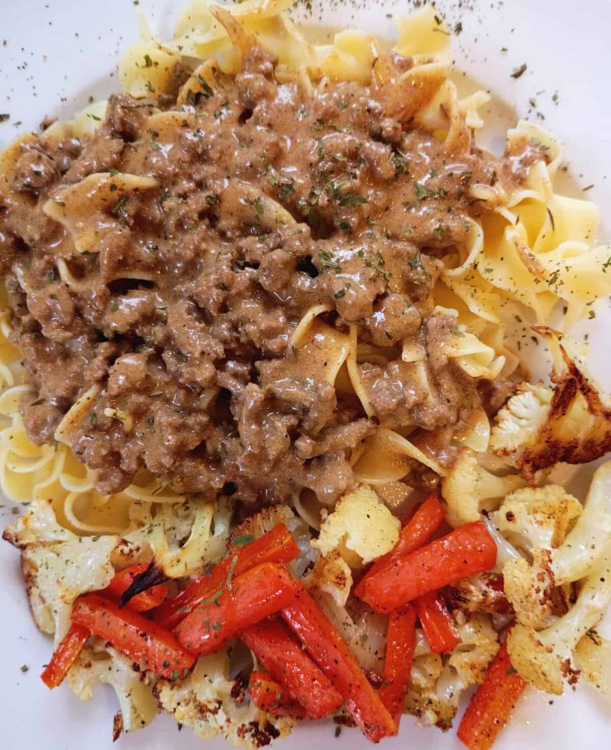 Pasta with creamy ground beef sauce, roasted carrots, and cauliflower, garnished with herbs on a white plate.