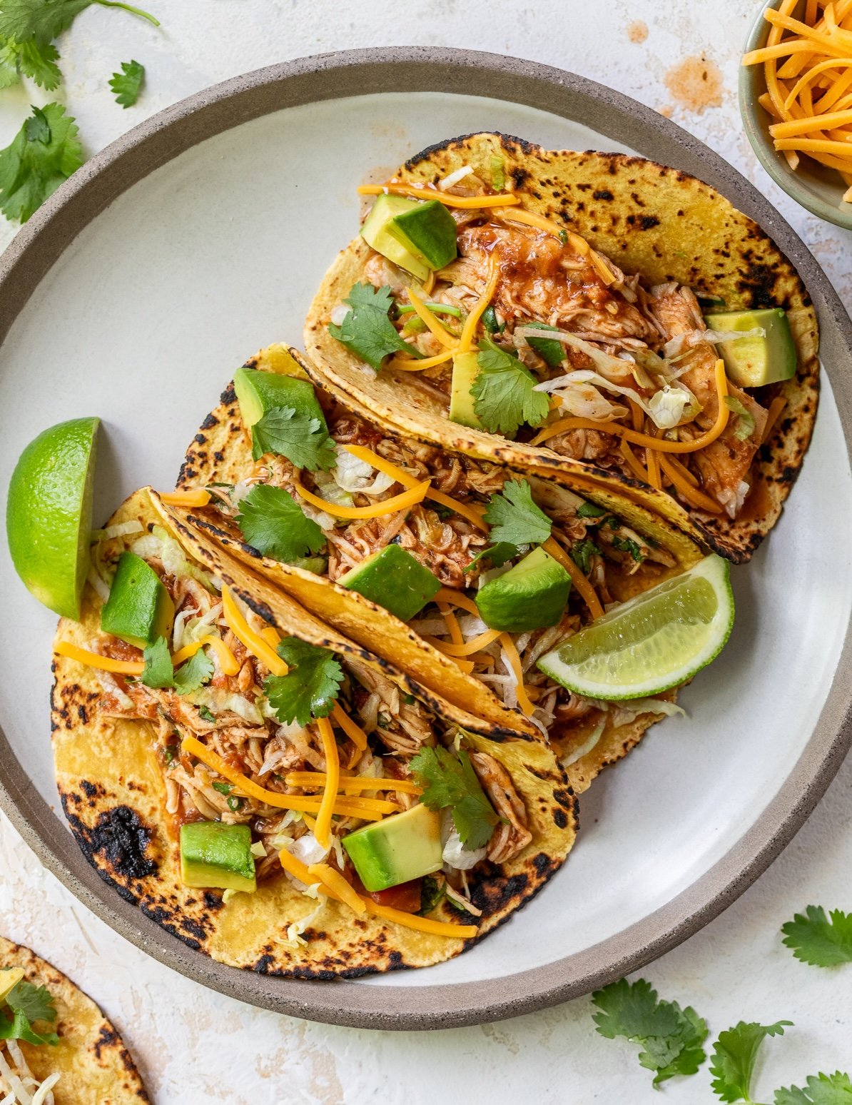 Three crockpot chicken tacos filled with shredded chicken, shredded cheese, avocado chunks, fresh cilantro, and lime wedges on a gray-rimmed plate. Some cilantro and food are scattered on the white surface nearby.