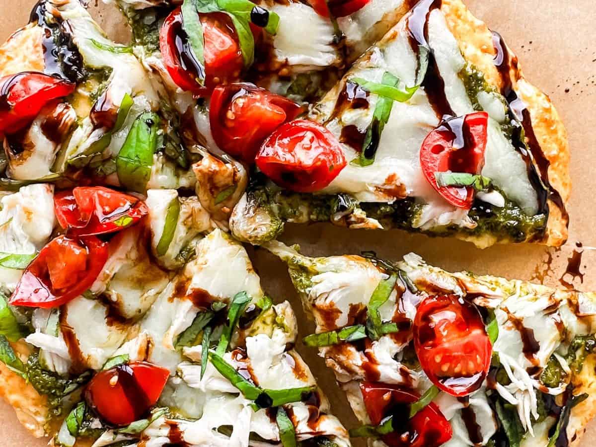 cottage cheese flatbread topped with melted cheese, fresh basil, sliced cherry tomatoes, and drizzled with balsamic glaze. Pieces are cut, revealing a crispy crust beneath the toppings. Placed on brown parchment paper.