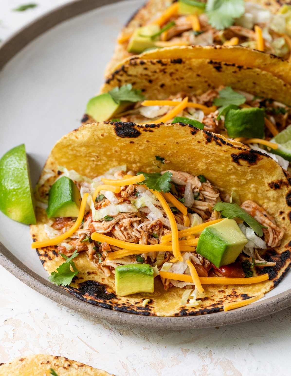 Three crockpot chicken tacos on a plate filled with shredded chicken, shredded cheddar cheese, chopped avocado, cilantro, and shredded lettuce, served in grilled corn tortillas with lime wedges on the side.