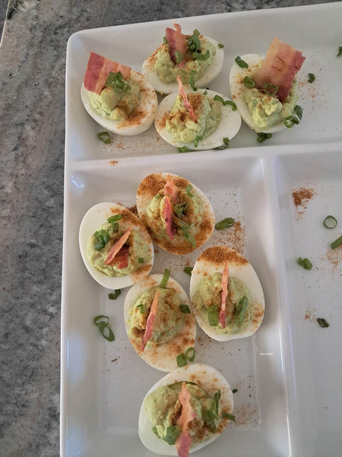 Deviled eggs topped with green filling, bacon strips, green onions, and seasoning on a white tray on a granite counter.