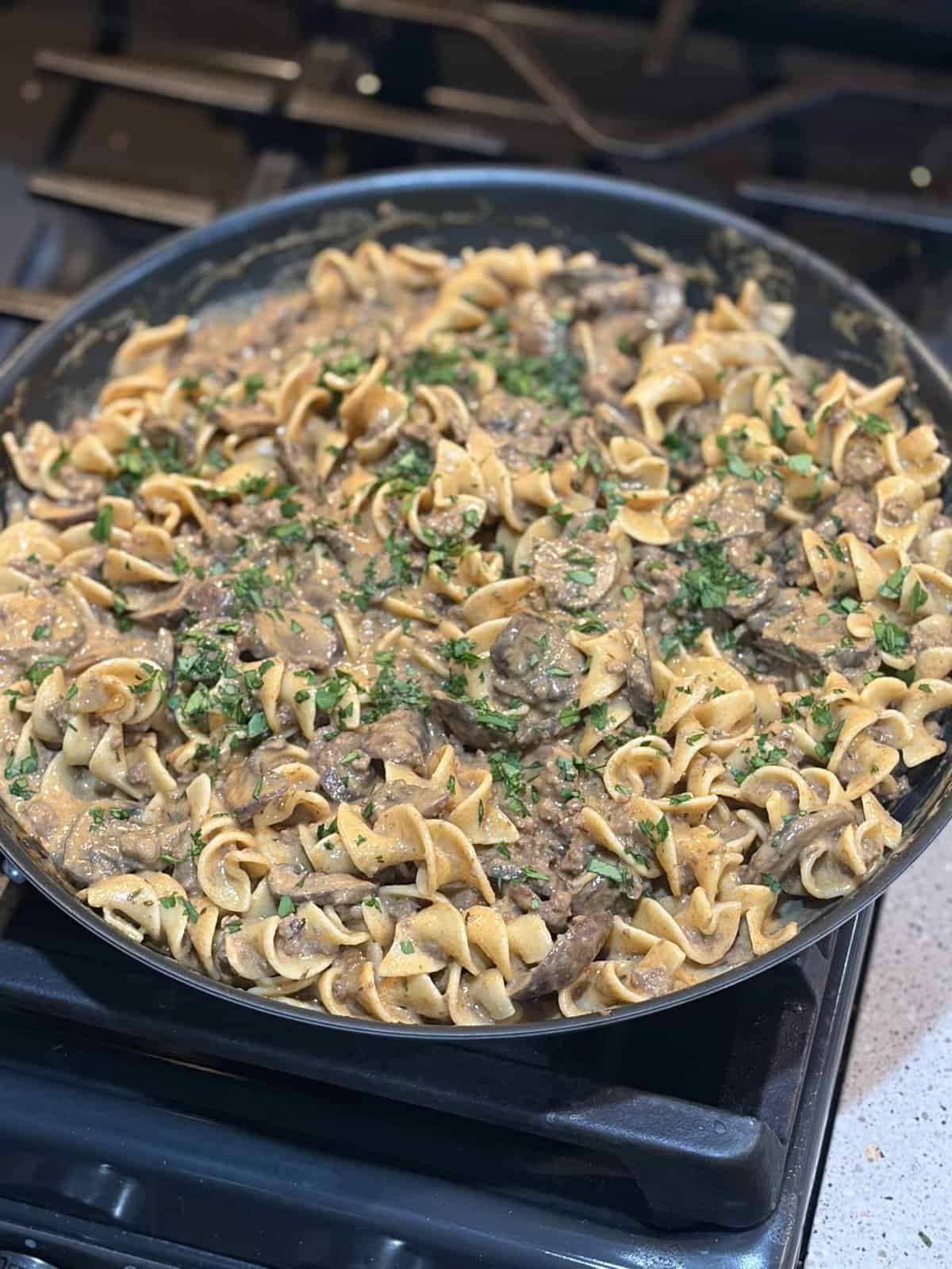 A skillet of creamy beef stroganoff with egg noodles and fresh herbs, sitting on a stove.