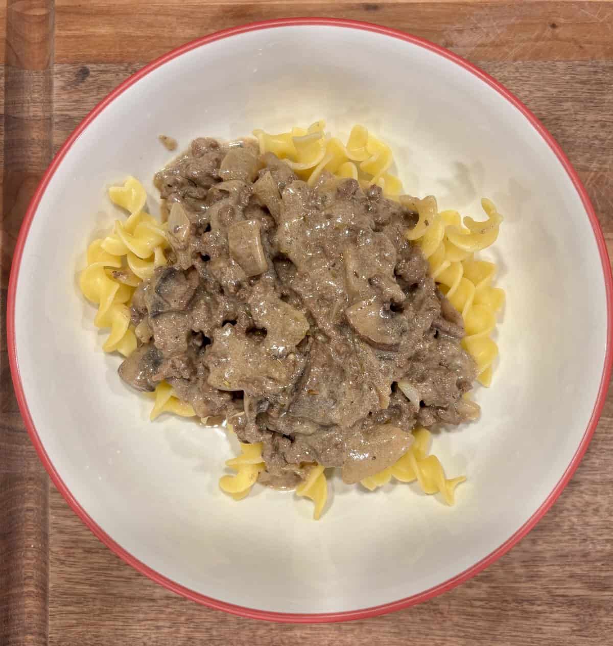 A bowl of egg noodles topped with creamy beef and onion stroganoff, on a wooden surface.