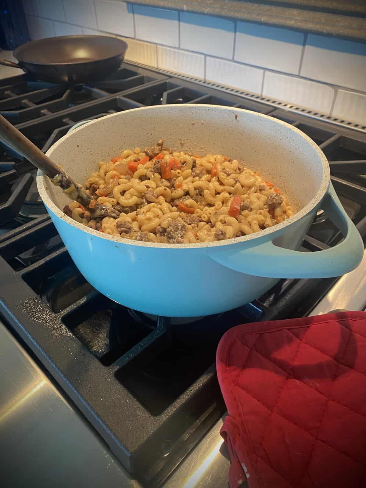 Large blue pot of pasta with meat and vegetables cooking on a stove, next to a red oven mitt.