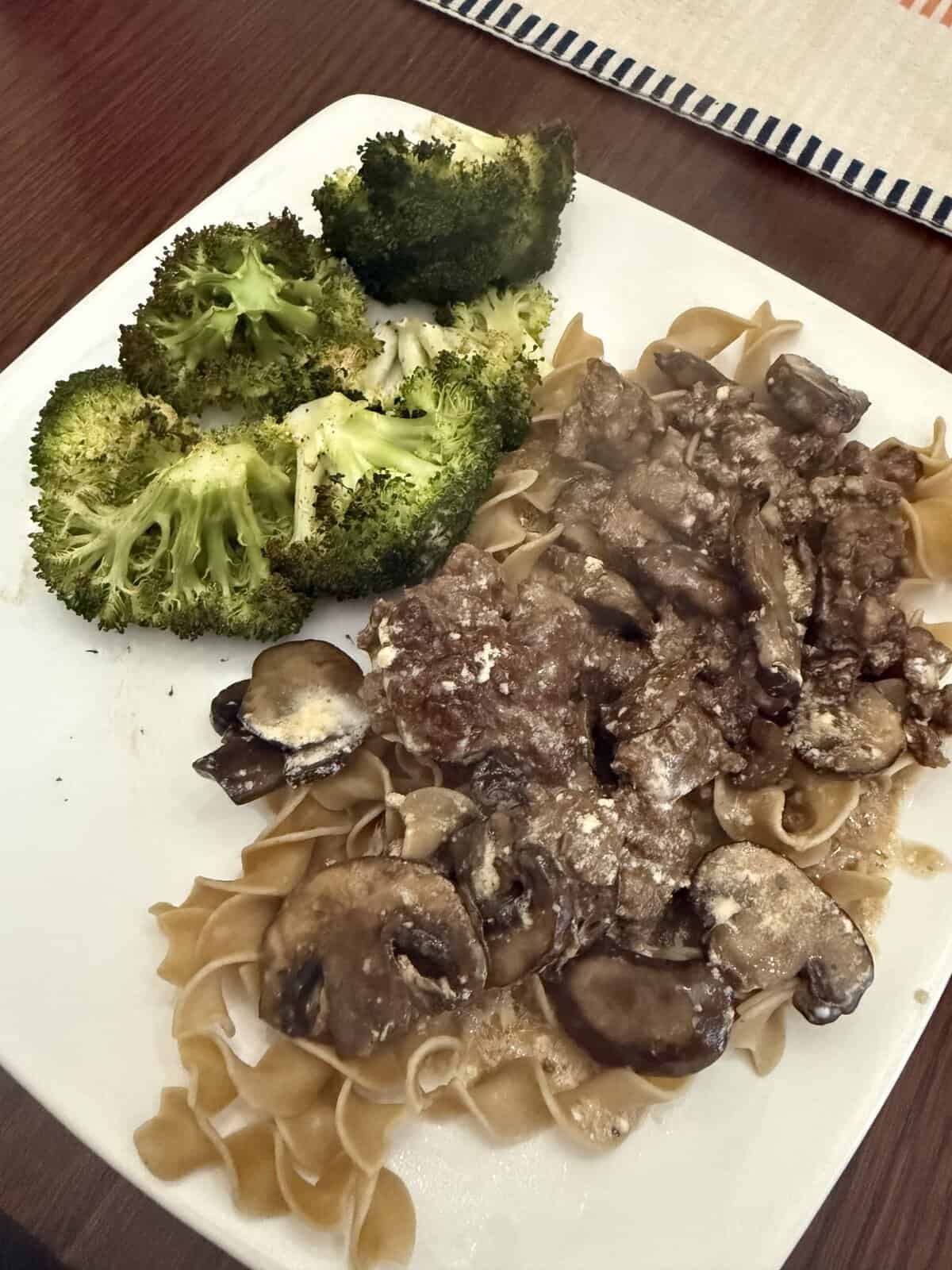 A plate with broccoli, egg noodles, and beef with a creamy mushroom sauce on a wooden table.