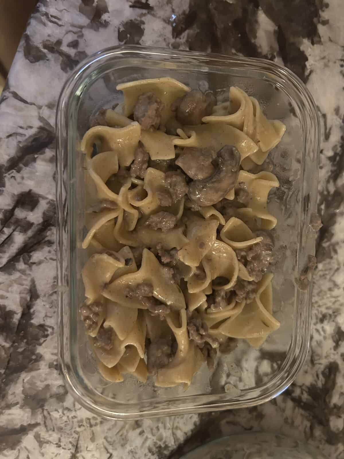 A glass container filled with egg noodles, ground beef, and mushrooms on a marble surface.