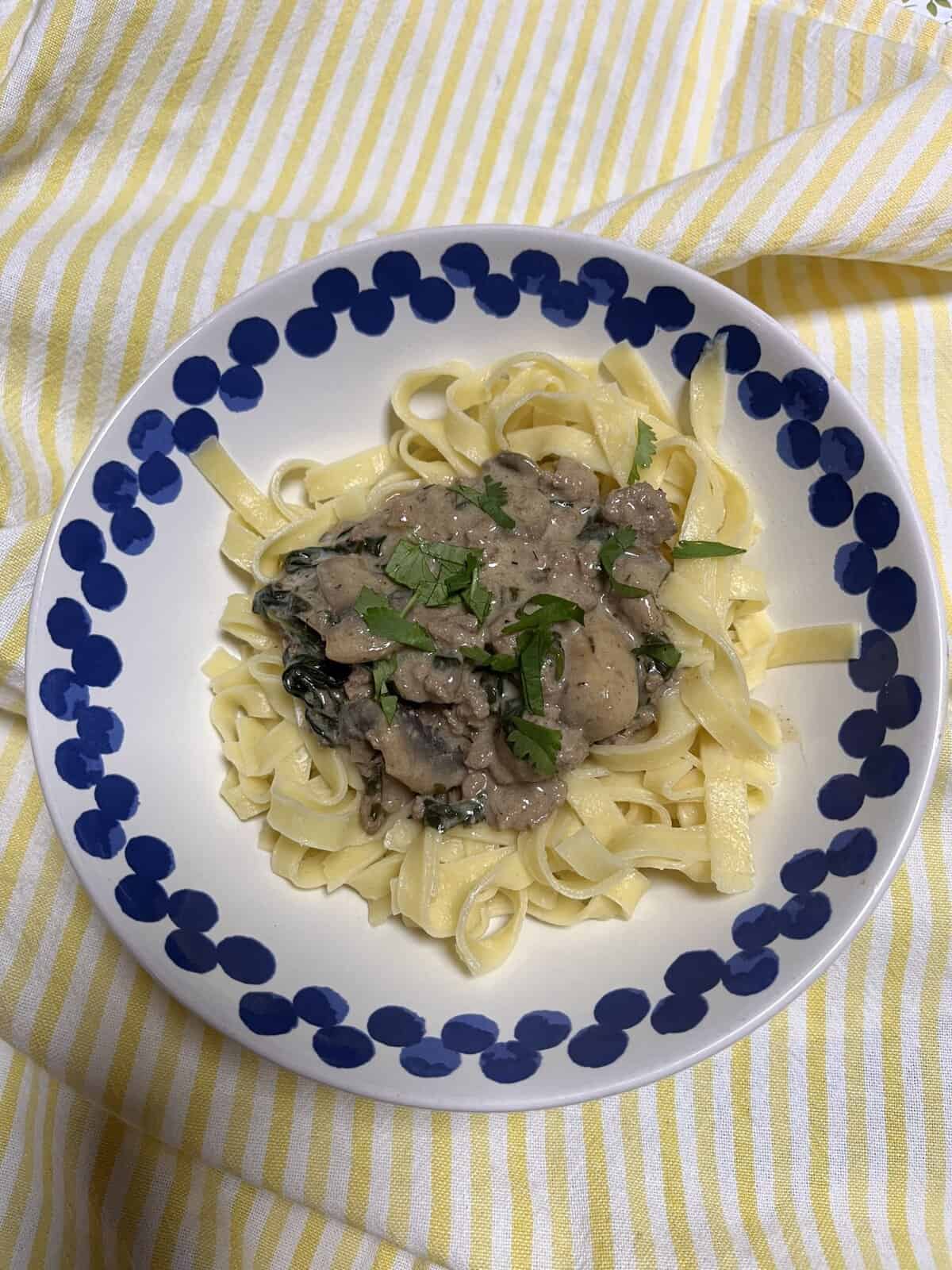 A plate of tagliatelle pasta topped with creamy mushroom sauce and herbs, on a blue-rimmed dish over a striped cloth.