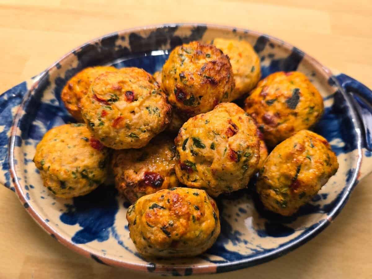 A ceramic dish holds several golden-brown, herb-filled baked meatballs on a wooden surface.
