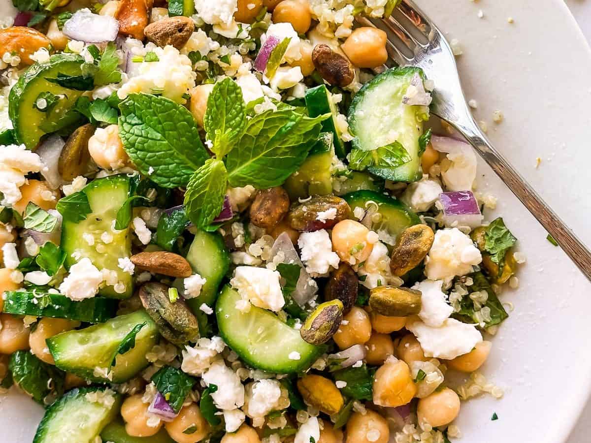 A fresh salad in a bowl featuring chickpeas, quinoa, cucumber slices, red onion, crumbled feta, pistachios, and mint leaves. A fork rests on the side of the bowl.