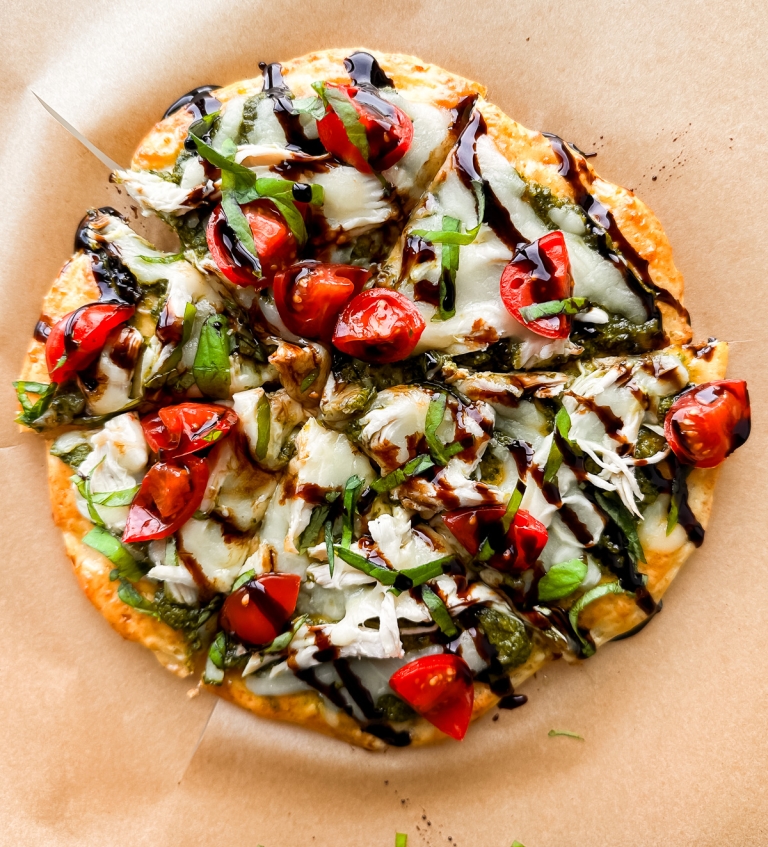 A rustic cottage cheese pizza topped with fresh basil, halved cherry tomatoes, and melted mozzarella cheese, drizzled with balsamic glaze. It's set on a piece of brown parchment paper.