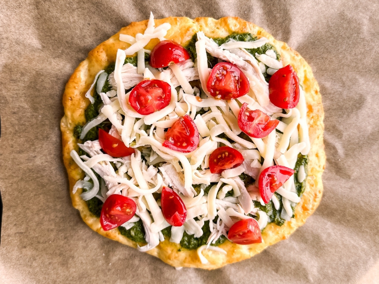 A homemade pizza with a cottage cheese crust on brown parchment paper. Topped with green pesto, shredded chicken, melted mozzarella cheese, and halved cherry tomatoes.