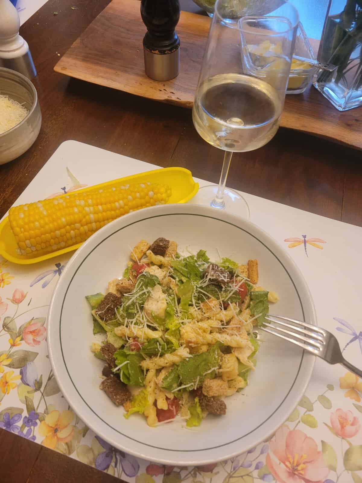 A bowl of salad, corn on the cob, and a glass of white wine on a floral placemat.